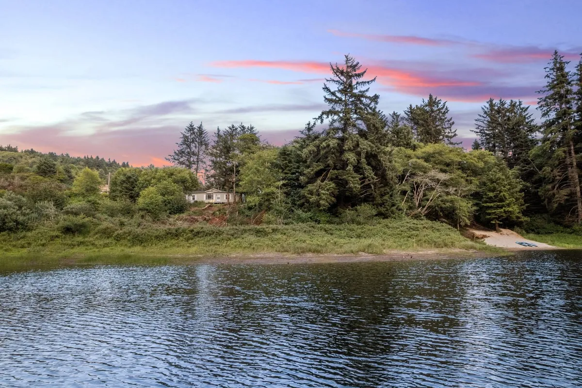 The house overlooks the lake at sunset, with trees framing the scene and the water reflecting golden light. Visible activities include fishing, boating, and strolling along the sandy beach, perfectly capturing a day winding down by the lakeside.