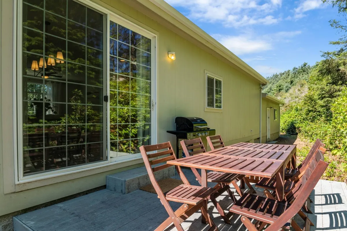 An inviting outdoor dining area with a wooden table and grill set against the backdrop of large glass windows, perfect for lakeside barbecues and group meals. The space exudes a casual and welcoming atmosphere ideal for gatherings.