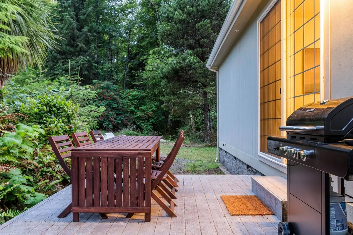 A cozy patio setup featuring a wooden dining table and a grill, enveloped by verdant foliage. The arrangement is perfect for barbecuing and hosting family celebrations in a relaxed setting.