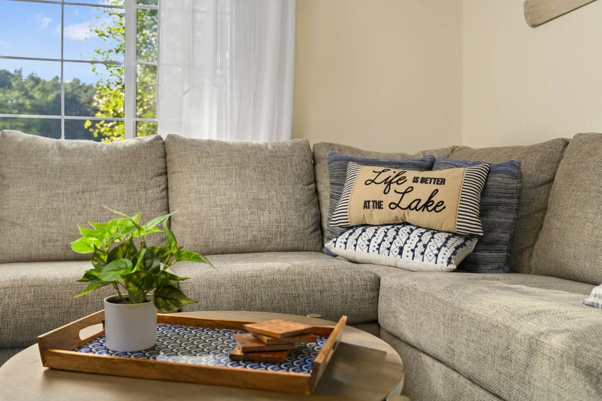 A cozy living area with a large, inviting sectional sofa adorned with decorative pillows. A coffee table with a tray sits at the center, and the room is filled with natural light from nearby windows, evoking a sense of comfort and spaciousness.
