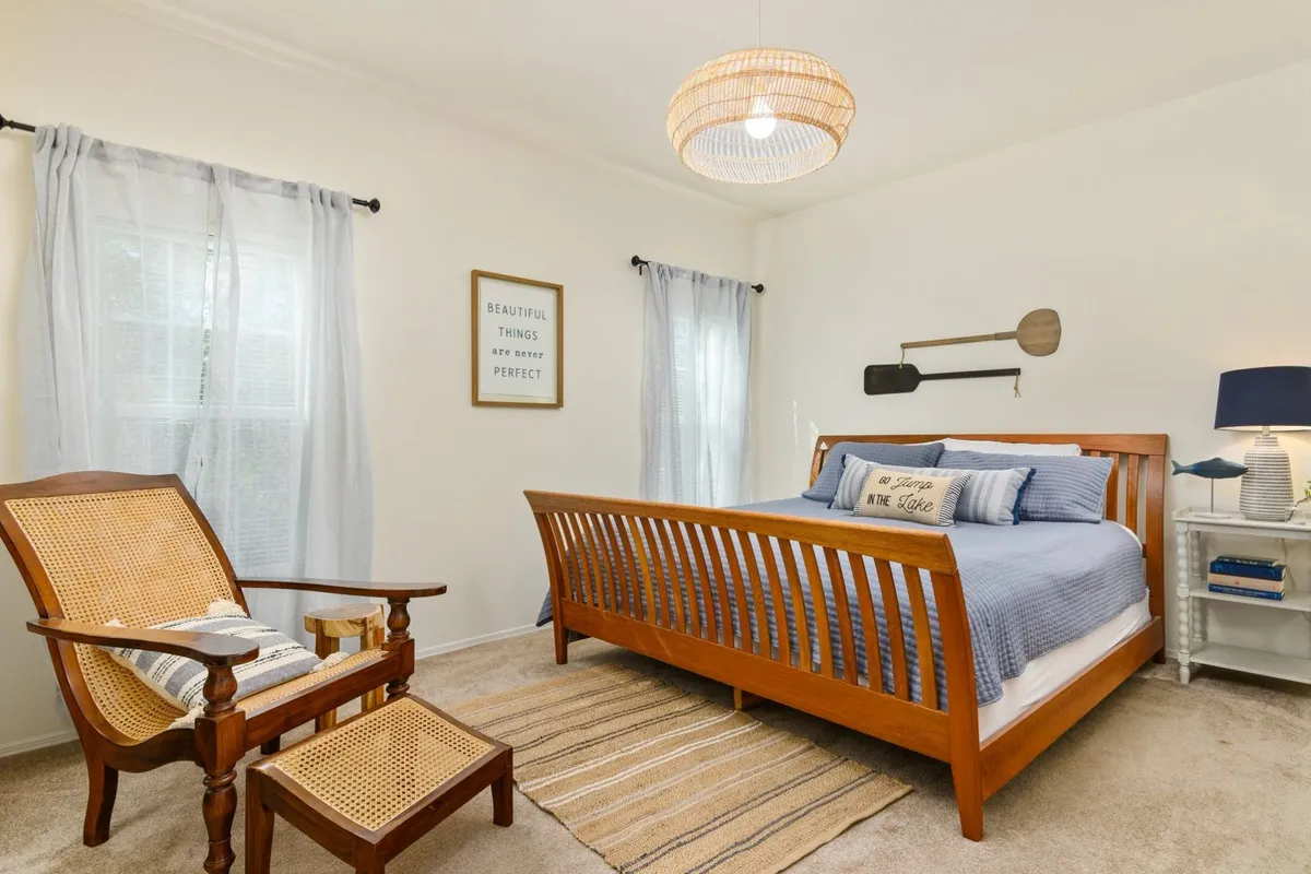 A serene bedroom featuring a wooden bed frame, woven chair, decorative wall art, and a bedside lamp. Sunlight streams gently into the room, highlighting the cozy and relaxing atmosphere, perfect for restful nights or quiet reading.