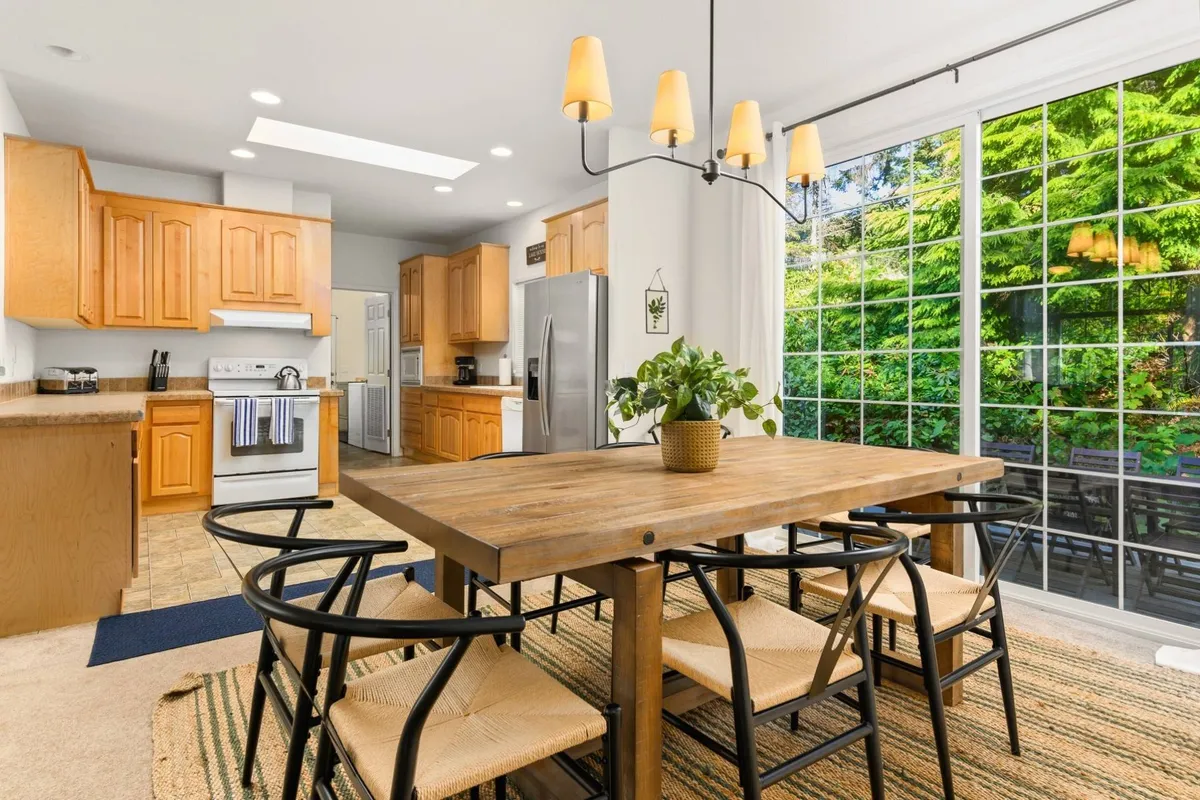A bright, open-concept living space featuring a central dining table, large windows that flood the area with natural light, and the adjoining kitchen with cabinetry visible in the background. The scene captures a sense of togetherness, with the layout flowing naturally from kitchen to dining to living areas, making it ideal for gatherings and shared meals.