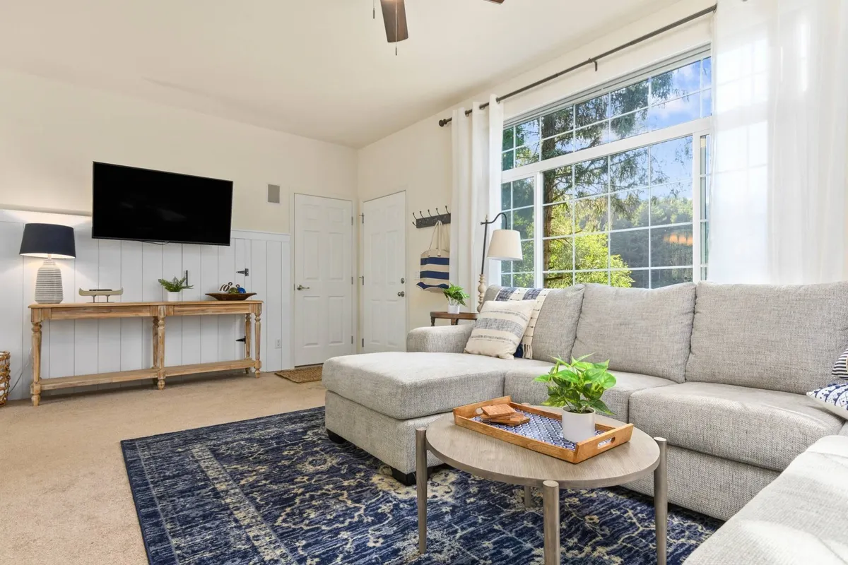 A comfortable sectional sofa positioned near expansive windows, decorative side table, and a television mounted on the wall. The space feels cozy and luminous, with sunlight streaming in, perfect for relaxing, playing games, or enjoying time together.