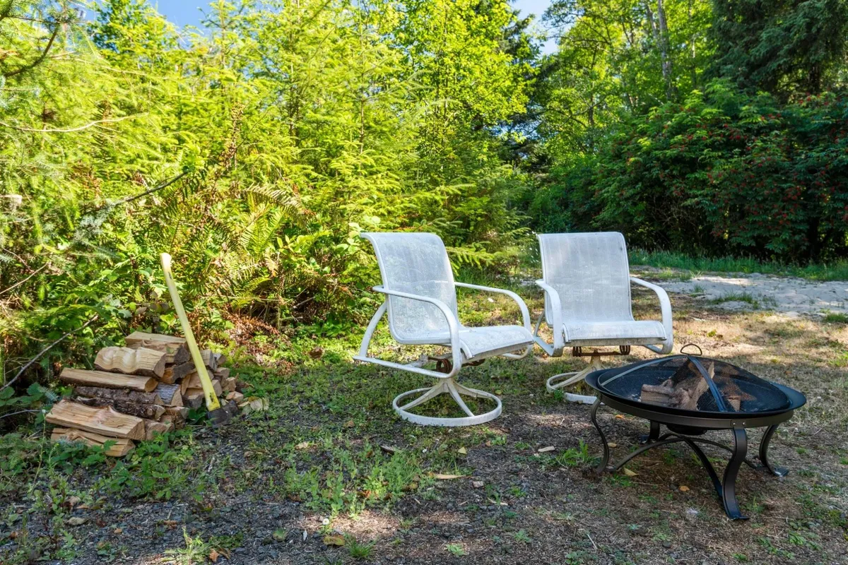 An inviting firepit scene with two white chairs and a tidy stack of firewood, surrounded by greenery. The mood is calm and perfect for stargazing or sharing stories by the fire.