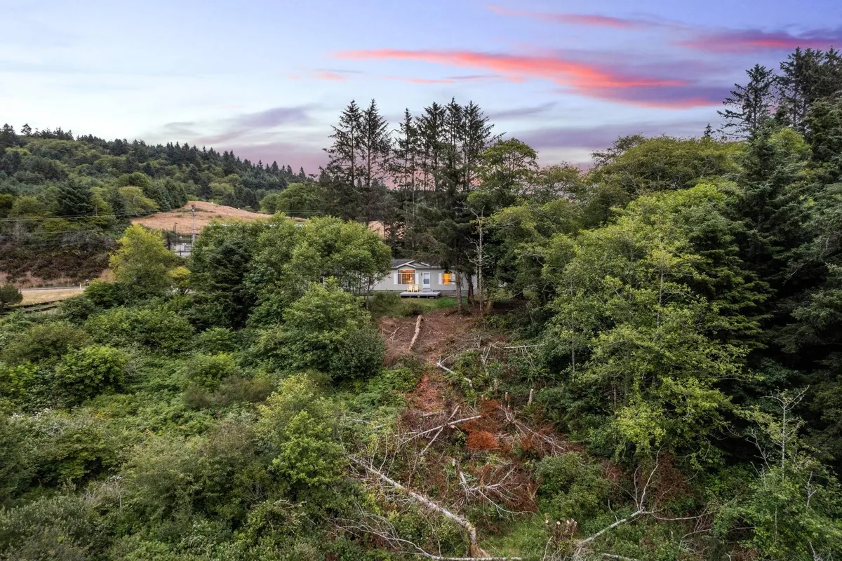 This image captures the house nestled among mature trees with a stunning sunset sky in the background. The view highlights the natural setting, with the home surrounded by greenery, evoking tranquility and connection to nature.
